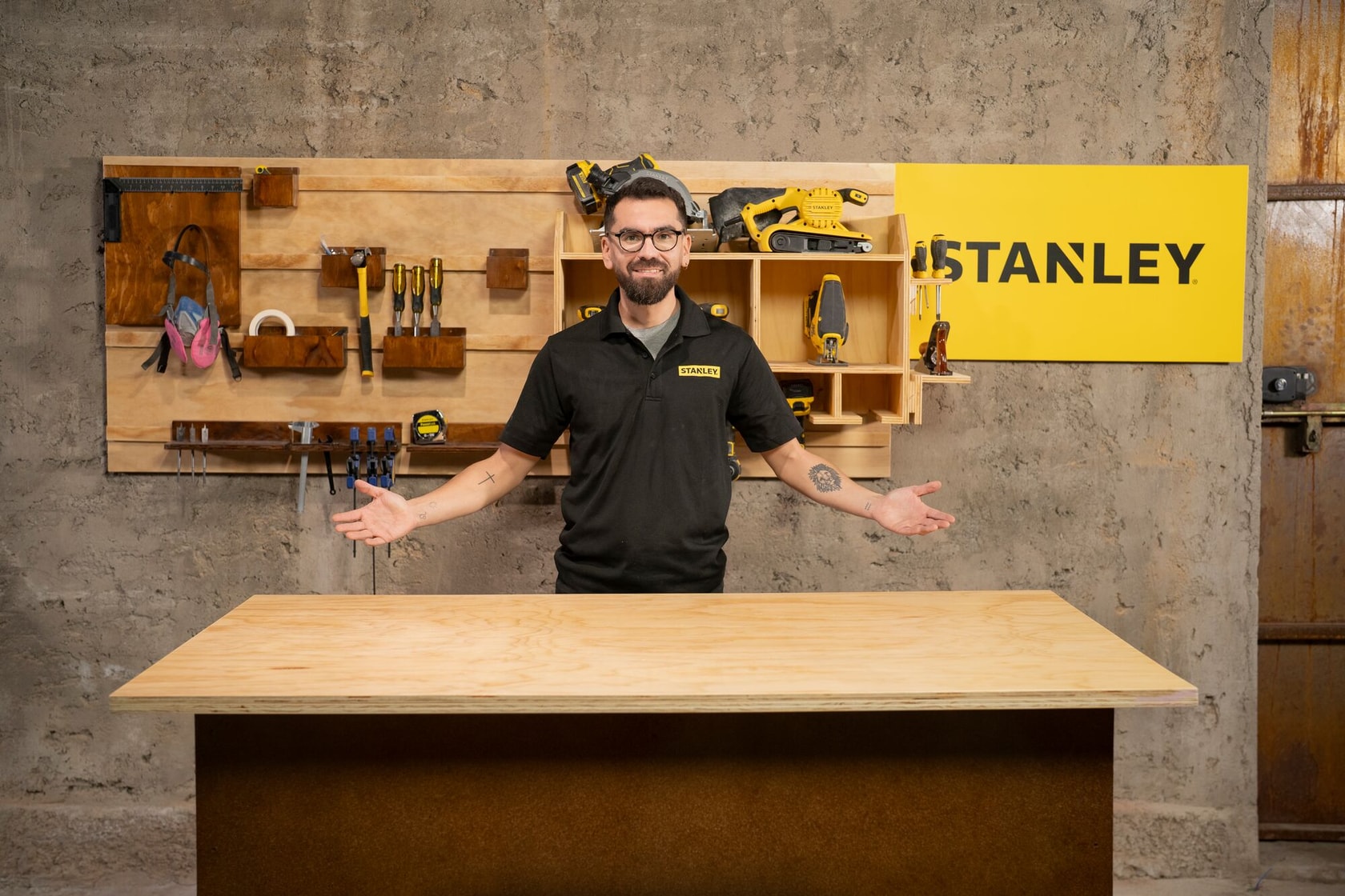 Una persona con camiseta negra de Stanley y gorra gris está de pie detrás de un banco de trabajo de madera en un taller. Al fondo, una pared con herramientas manuales, eléctricas y un cartel amarillo de Stanley.