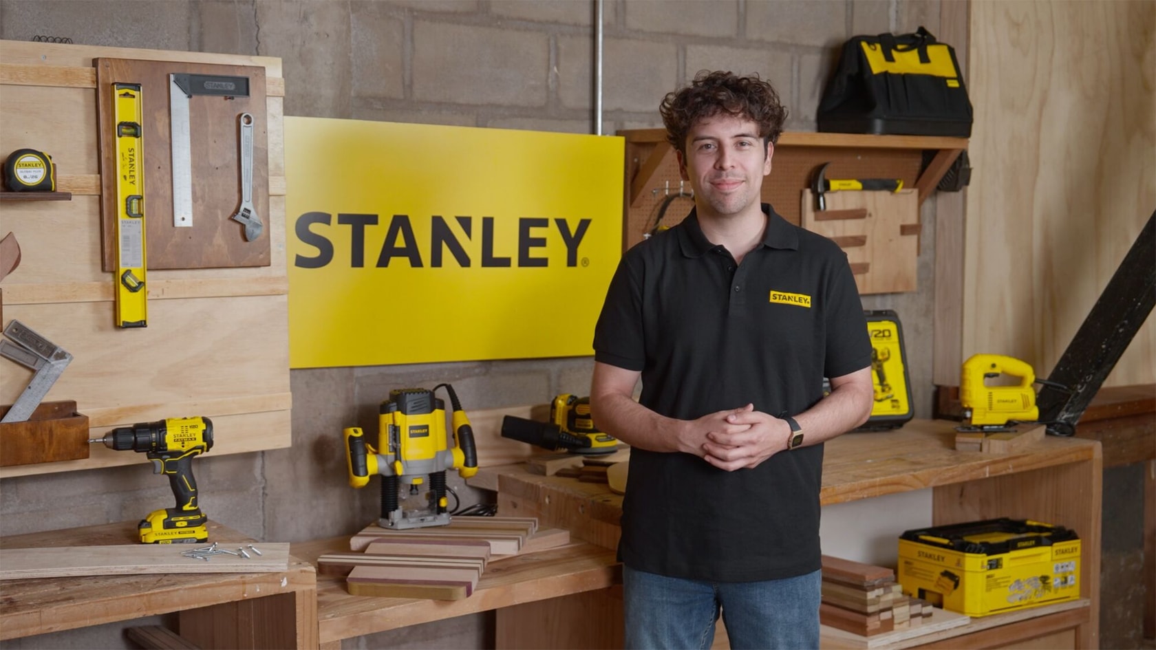 Una persona con el rostro difuminado está de pie en un taller frente a un cartel amarillo de STANLEY. Hay varias herramientas y equipos de la marca STANLEY, como un taladro, una cinta métrica, un nivel, una bolsa de herramientas y cajas, organizados en bancos y estantes de madera.
