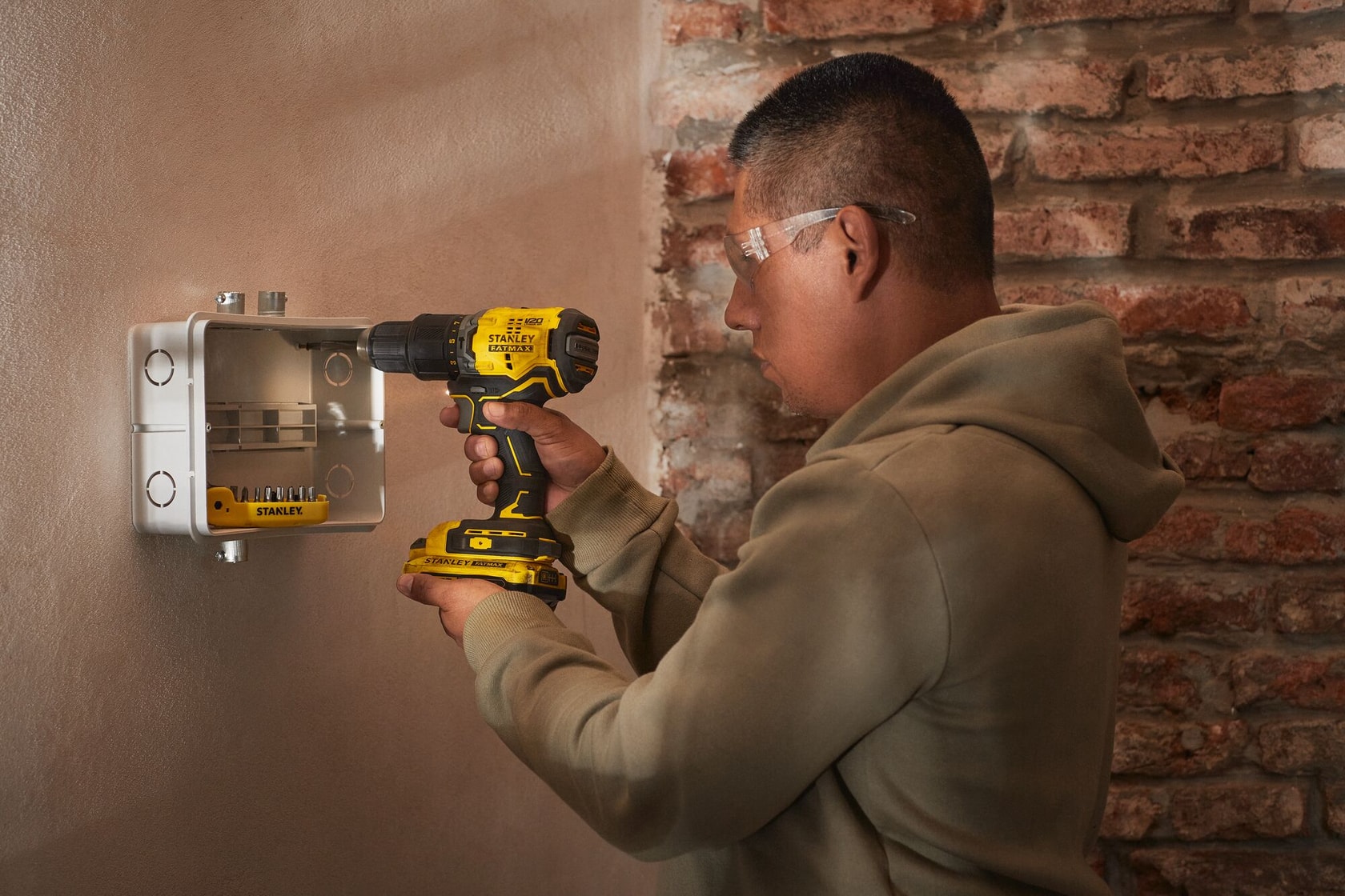 Foto de persona usando taladro inalámbrico STANLEY en caja metálica de pared, usando gafas de seguridad.