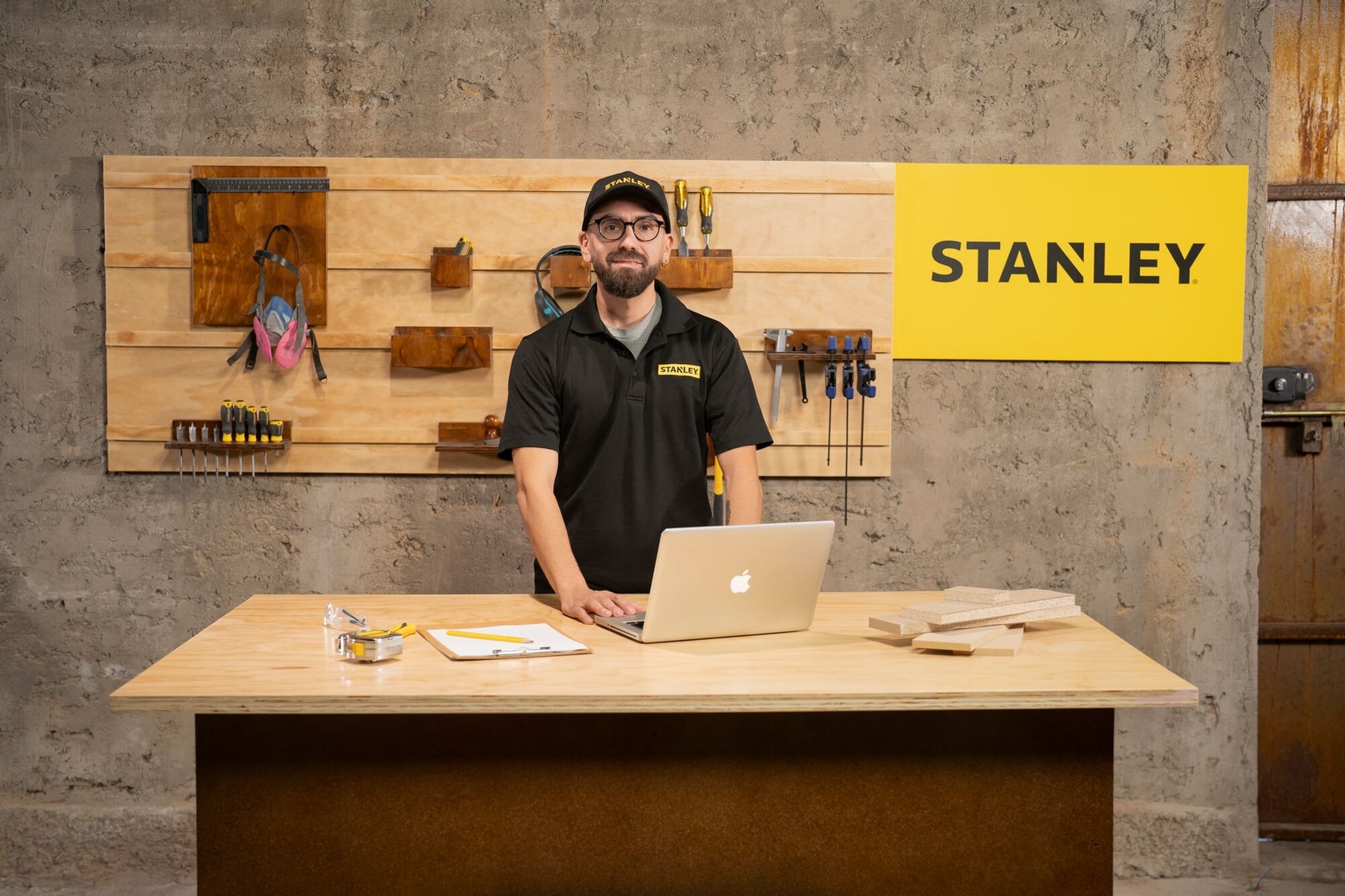 Una persona con camiseta y gorra negras de la marca STANLEY está de pie detrás de una mesa de trabajo de madera con un portátil, una carpeta, una cinta métrica y piezas de madera, en un taller. En la pared hay varias herramientas manuales y un gran cartel amarillo de STANLEY.