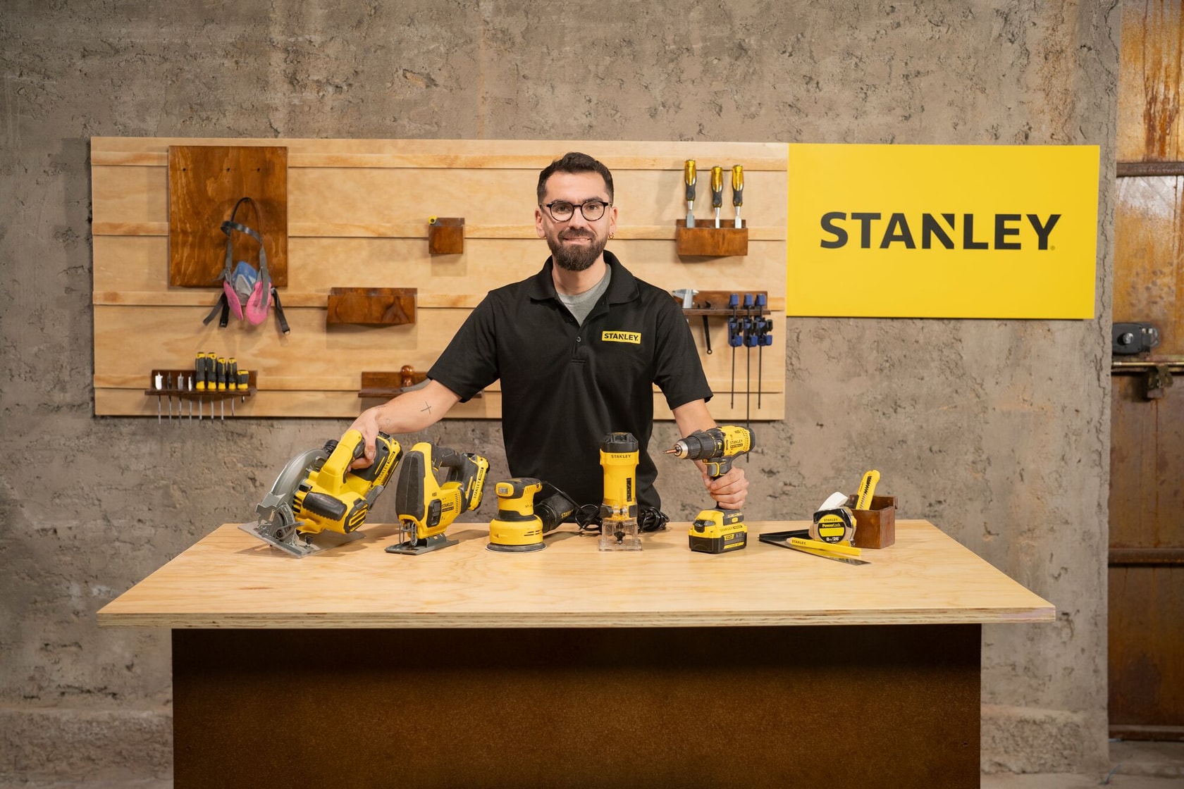 Una persona con camiseta negra STANLEY está de pie detrás de una mesa de trabajo de madera mostrando varias herramientas eléctricas STANLEY amarillas y negras en un taller. El fondo tiene un panel organizador de herramientas y un cartel amarillo de STANLEY.